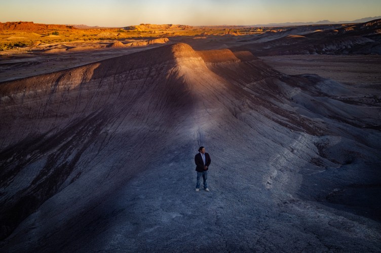 A wide, elevated view of a man standing alone on a vast, eroded gray ridge in a desert landscape. The setting sun casts a warm, golden glow across the tops of the distant hills.
