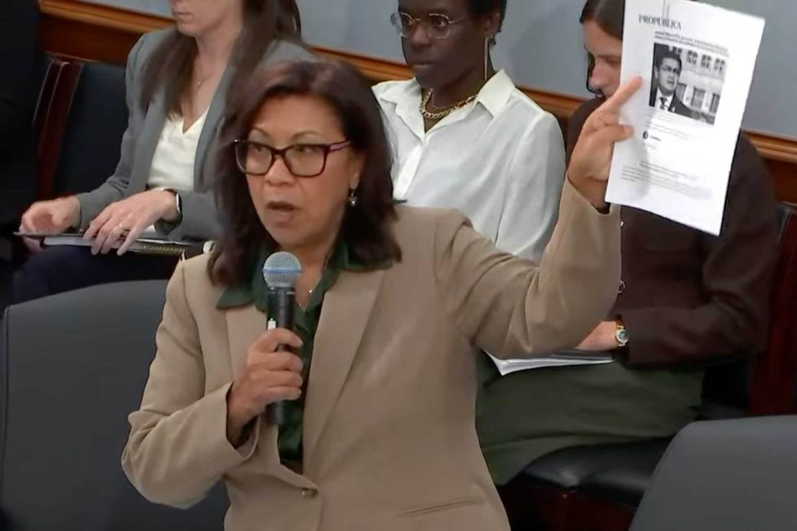 A woman wearing glasses and a tan blazer speaks into a handheld microphone while holding up a document featuring the ProPublica logo and a man's photograph. Several observers sitting in a row behind her, listening.
