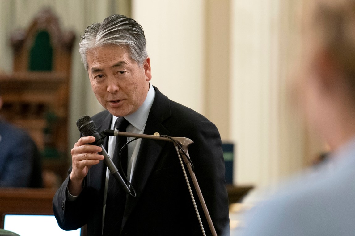 A man in a suit holds a microphone as he speaks in a government building.