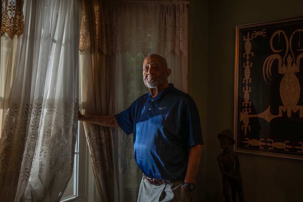 An elderly Black man stands in a dark room, looking at the camera. Window light illuminates one side of his face.