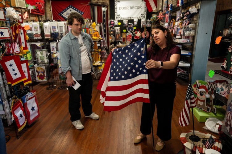 Two people stand in a room filled with U.S. flags and other decorations. One person holds a U.S. flag and considers it, and the other person looks on.