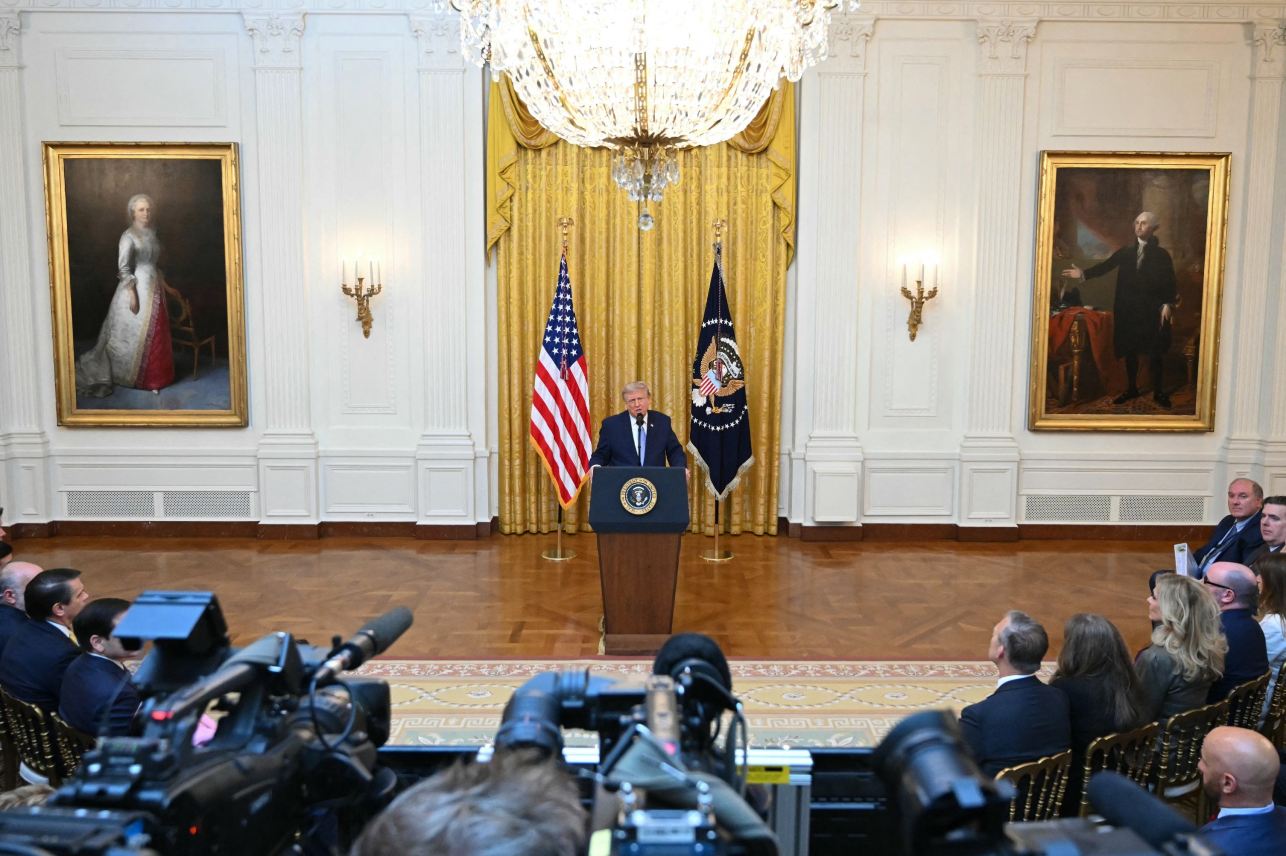 President Donald Trump stands at a podium in the East Room of the White House, with the U.S. flag, the presidential flag and a golden curtain behind him. On either side of him are portraits of Martha and George Washington, in golden frames. In the foreground are TV and news cameras and a seated audience.