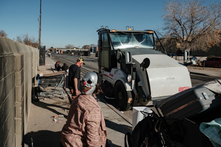 A street sweeper drives past two individuals as they pick up their belongings on the side of a road.