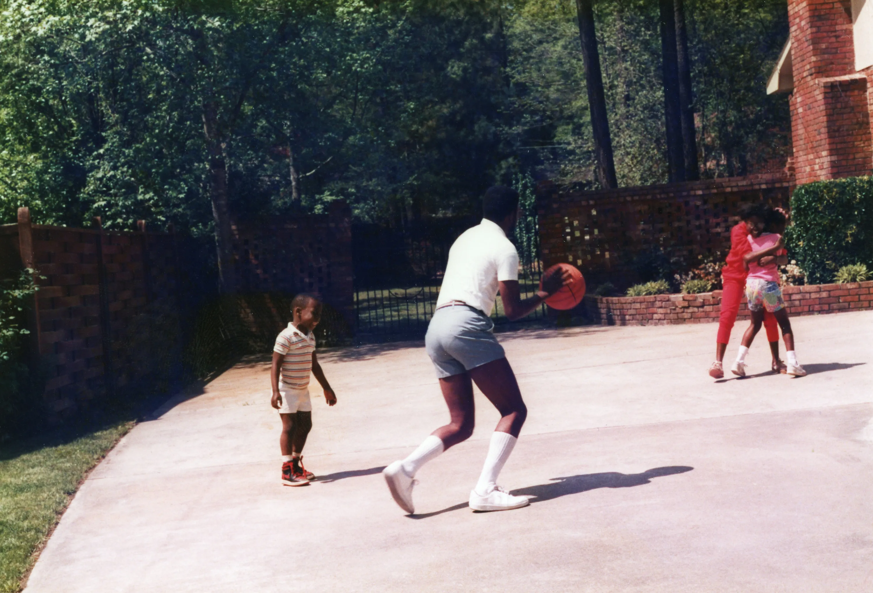 A man wearing shorts, long white socks and sneakers faces away from the camera holding a basketball. He is midaction while one child looks on. Two kids in the background are wrestling.