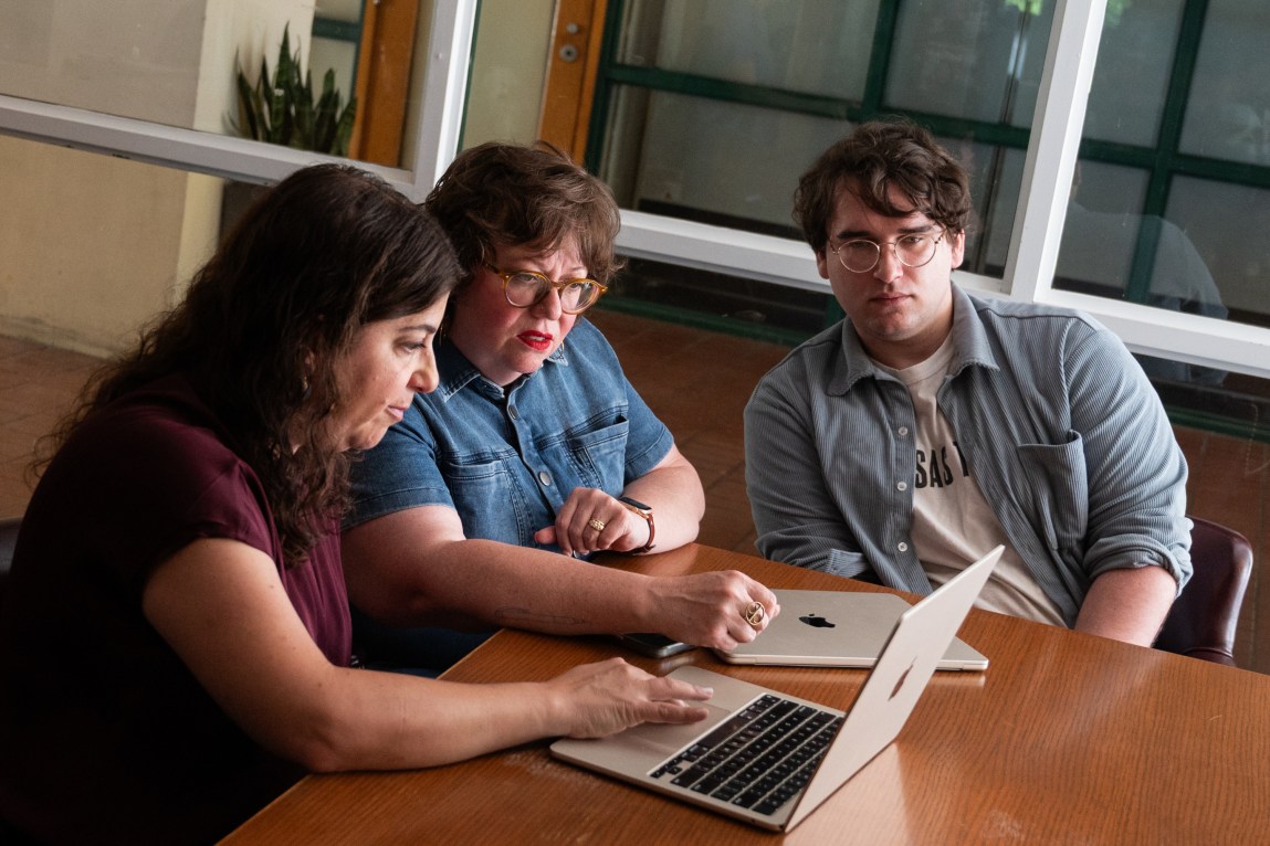 A group of three people sit in leather chairs at a wooden table, intently looking at an open computer. A second laptop sits on the desk. The room has glass windows.