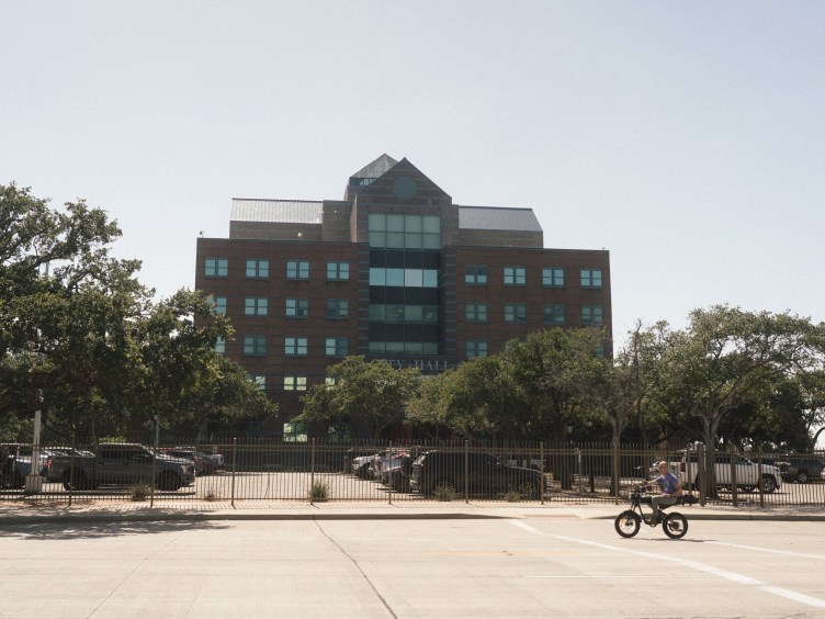 A wide shot shows a multistory brick building labeled "City Hall" behind a fenced-in parking lot. In the foreground, a person rides a black bicycle across the wide, paved street under a bright clear sky.