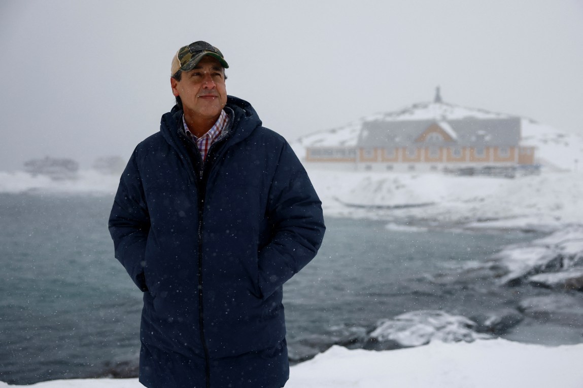 A man wearing a baseball cap and long blue coat stands in front of a body of water, a snowy landmass and an out of focus building in the distance.