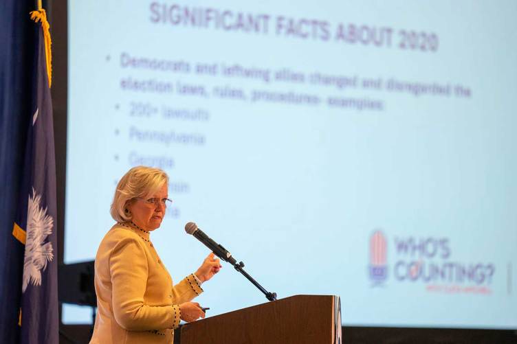 A woman gestures from behind a wooden lectern as she speaks into a microphone. She is standing beside a South Carolina state flag with a presentation slide titled “SIGNIFICANT FACTS ABOUT 2020” projected behind her.