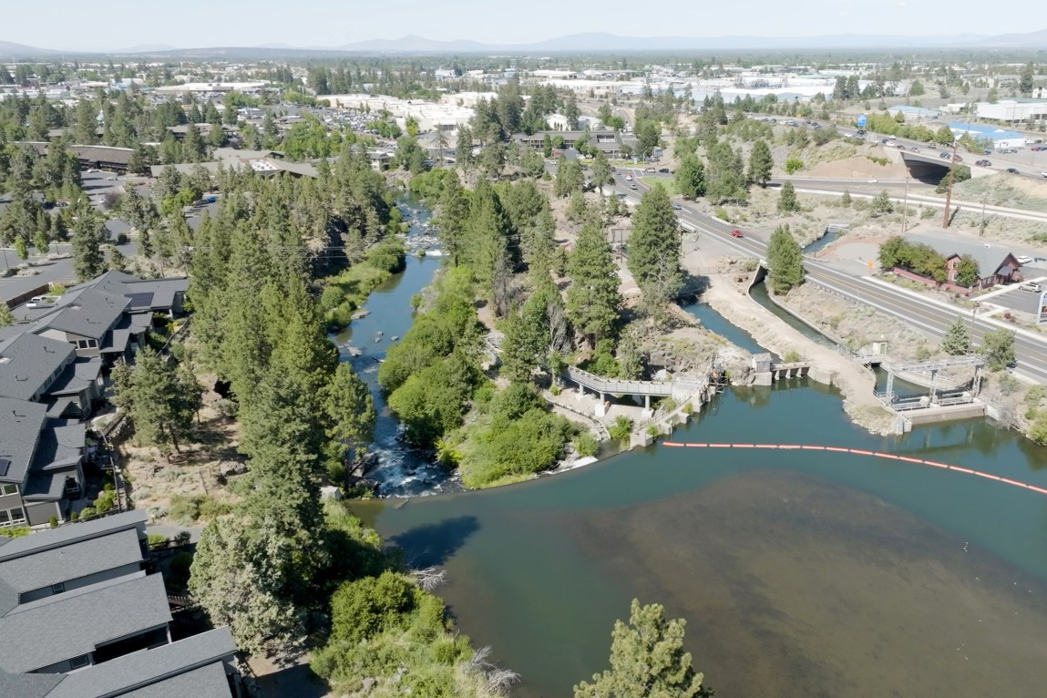 A river, seen from above, flows into a dam, and then emerges without 90% of its water. A row of houses overlooks the river on the left side, and a highway runs alongside it on the right.