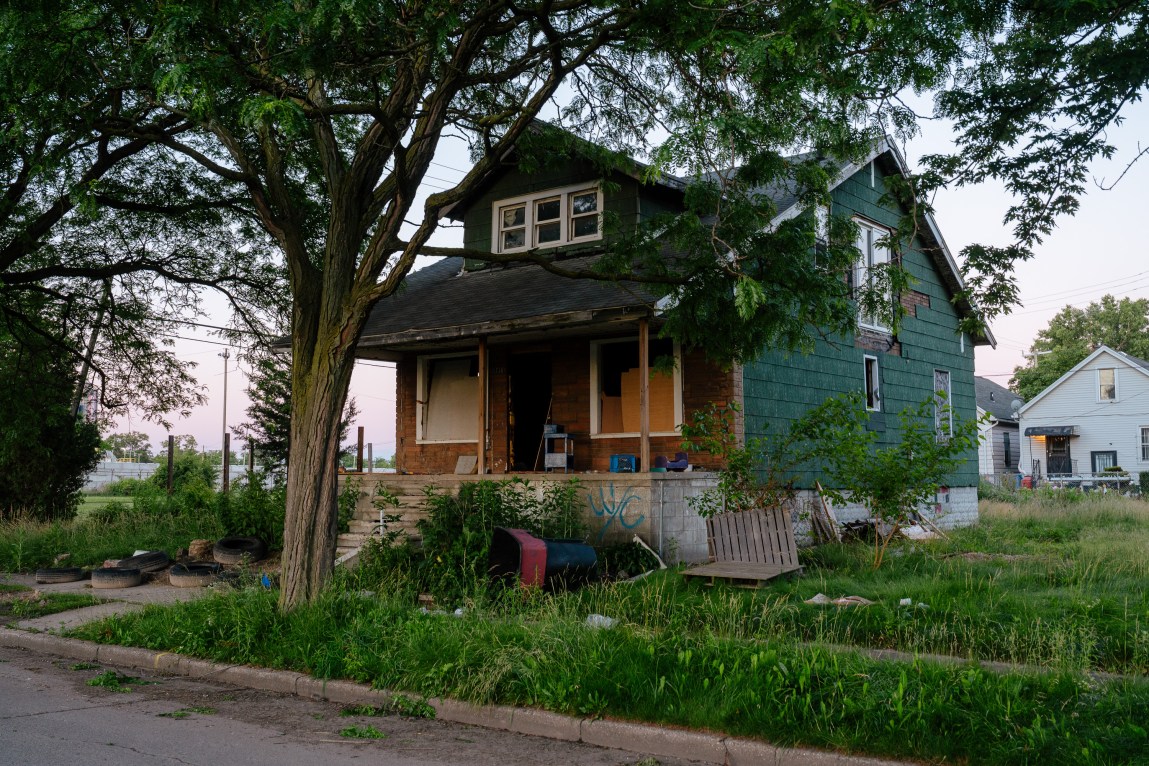 A dilapidated house with no front door or front windows and an overgrown, trash-covered lawn.