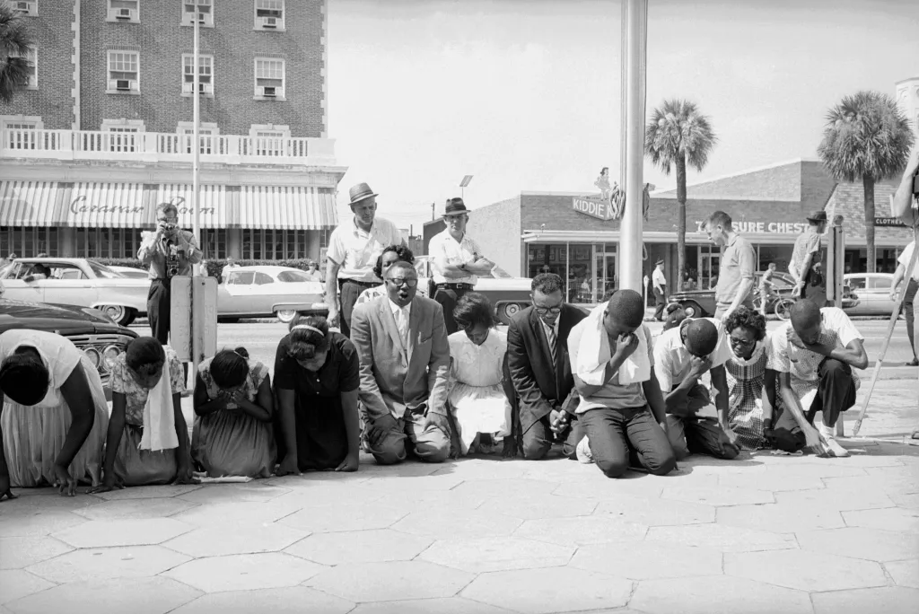On a busy street, a group of men and women kneel on the ground looking down at the sidewalk. A man in the center is wearing a suit and speaking with his eyes closed. There is a photographer and various men wearing hats lingering behind the group.