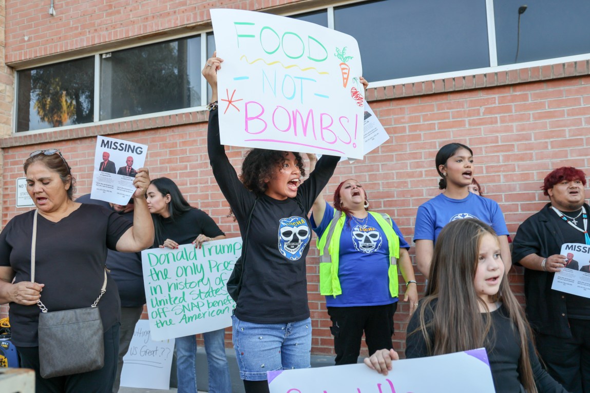 A woman with black hair, wearing a black, long-sleeve T-shirt with a screenprint of a lucha libre mark on the front, and blue jean shorts, holds up a sign with the slogan “FOOD - NOT - BOMBS” amid a crowd of protesters, some also carrying signs, in front of a red brick building with windows.