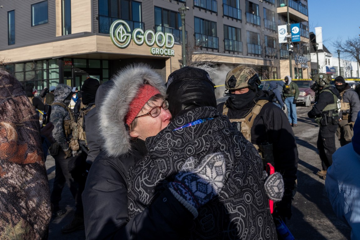 A woman in a coat with a furry trim on the hood holds someone in a hat and dark jacket. Behind them are clusters of men in tactical vests, some with helmets, many with masks on.