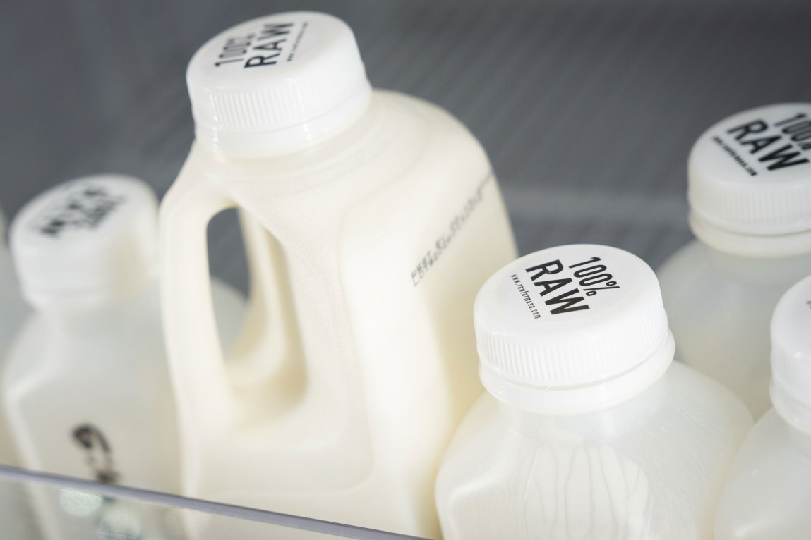 Milk bottles sit on a refrigerator shelf, sealed with caps displaying the text “100% RAW.”
