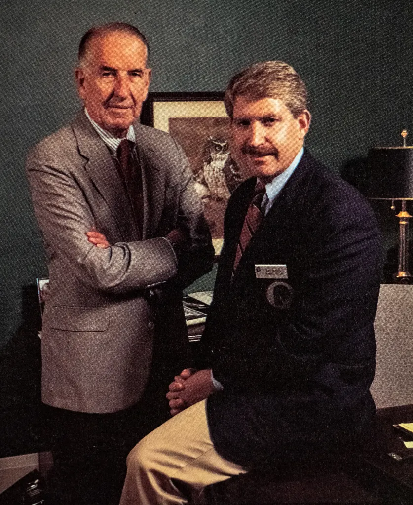 Two men with gray hair wearing suits pose together in an office.
