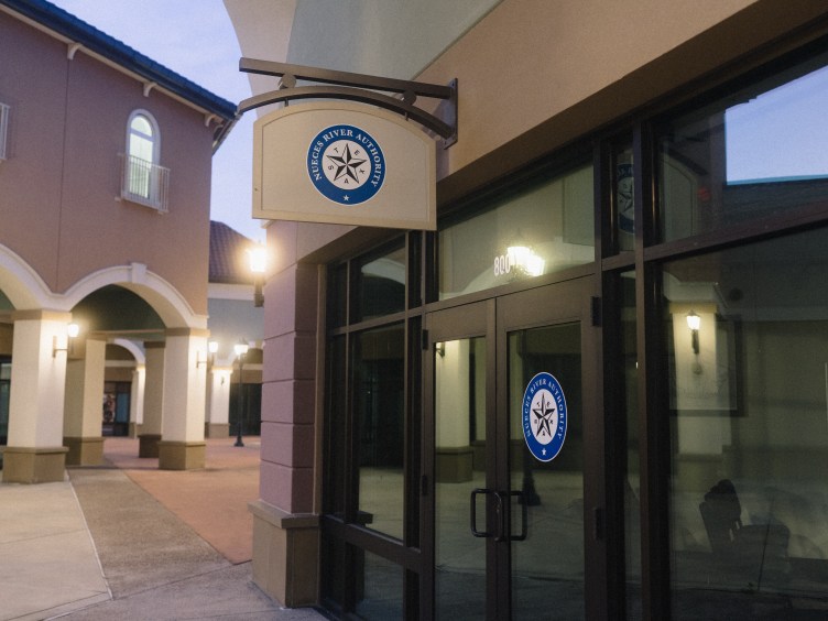 An exterior view of an office storefront featuring a sign with the Nueces River Authority logo — a blue circle surrounding a Texas star. 