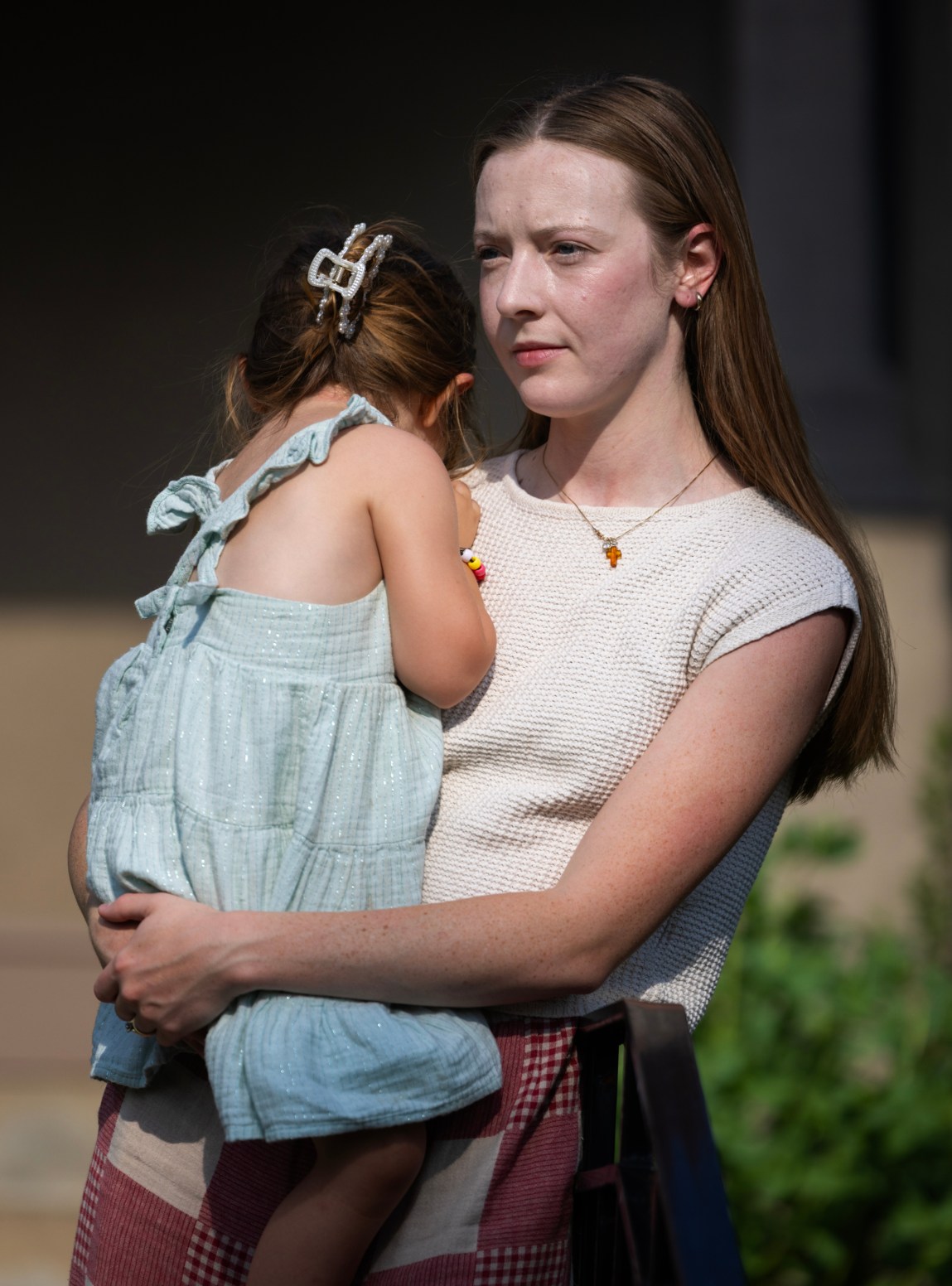 A woman with long auburn hair wearing a white shirt, checkered pants and a necklace with a cross holds a toddler turned away from the camera wearing a blue dress, beaded bracelet and hairclip.