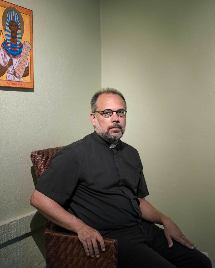 A man with a short beard and glasses, dressed in a black clerical shirt with a white collar, sits in a woven armchair looking directly at the camera. 