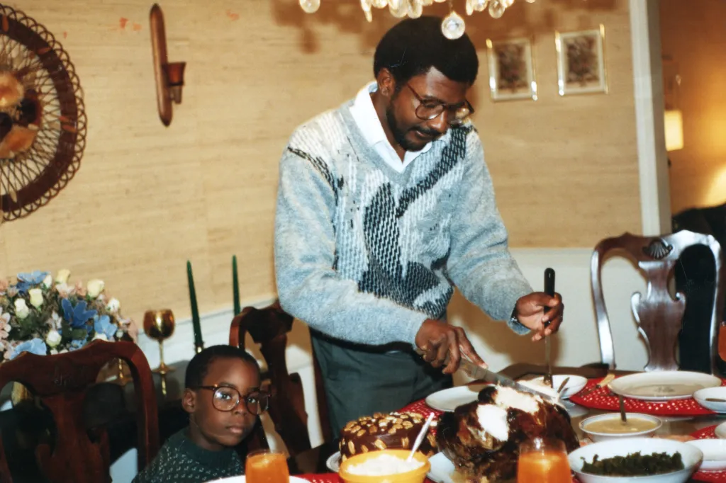 A homey room where a little boy wearing glasses sits at a table next to his father, who is wearing a V-neck sweater and glasses. The father is carving meat on a table full of dishes of food.