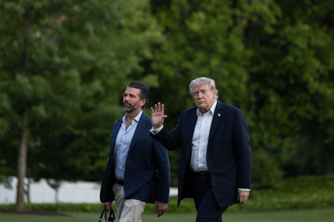 President Donald Trump waves and Donald Trump Jr. walks next to him. They are both wearing blazers and button-down shirts and have serious expressions.