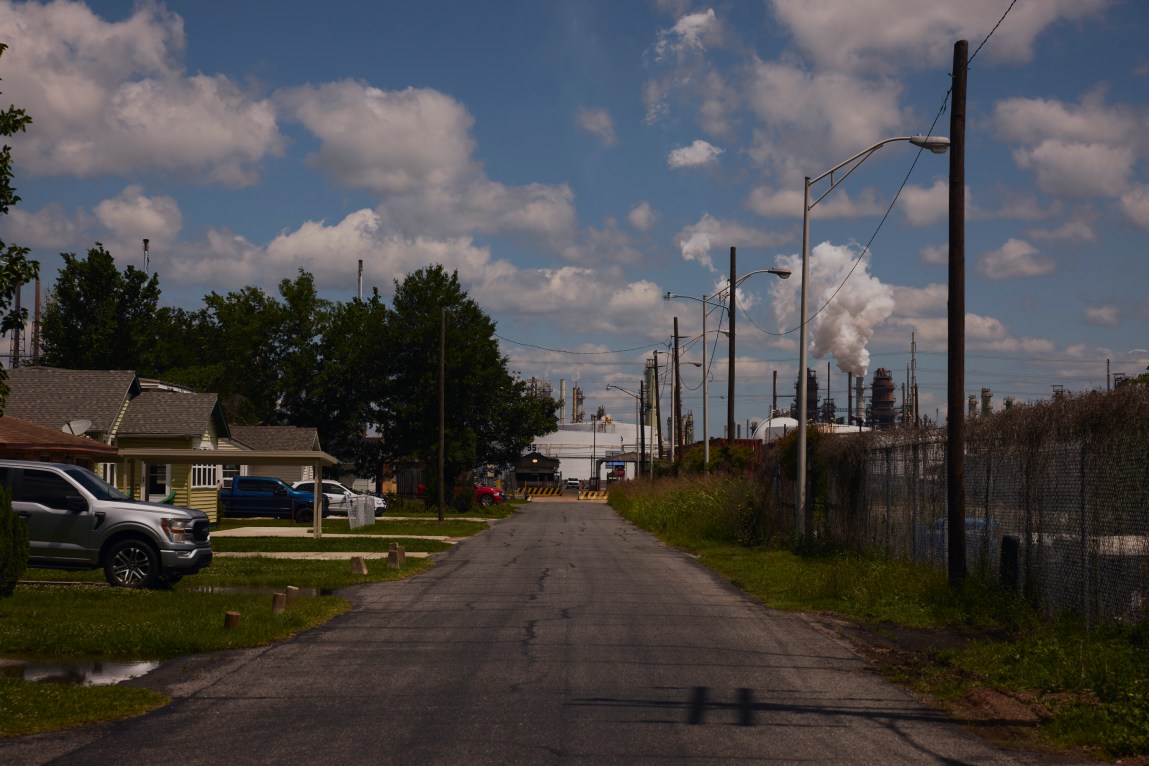 A street view looking down a road in a neighborhood, with houses on the left and an industrial facility with smokestacks emitting white clouds of smoke on the right, all under a bright blue sky with scattered clouds.