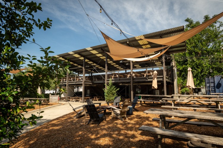 A patio with woodchips, Adirondack chairs, picnic tables, umbrellas, trees and shade structures.