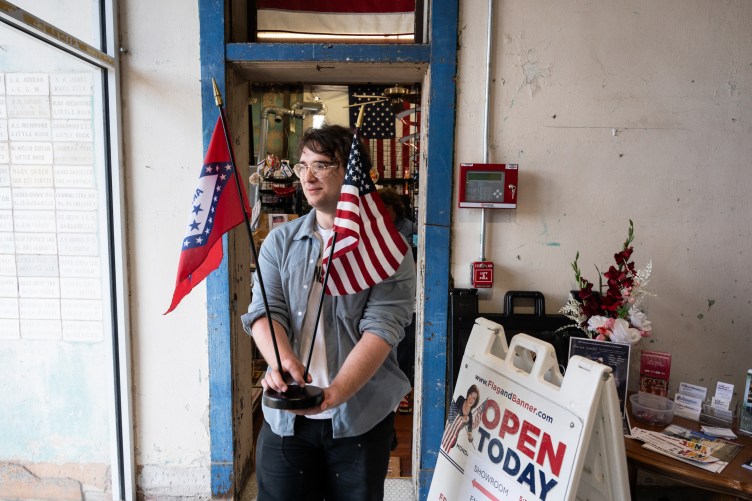 A person smiles and walks out of a blue doorway holding a stand with a small U.S. flag and Arkansas state flag.