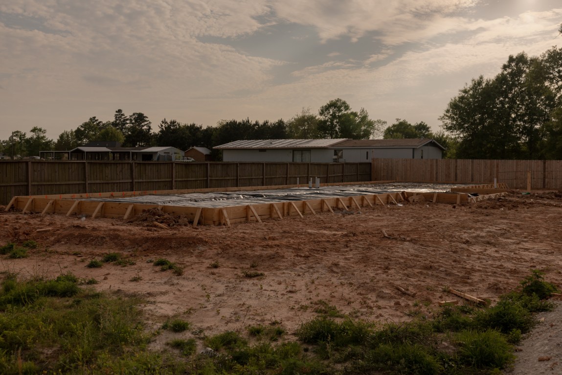 An unfinished foundation for a building, held up by wooden bracing in a muddy housing lot, surrounded by a wooden fence with houses and trees in the background.