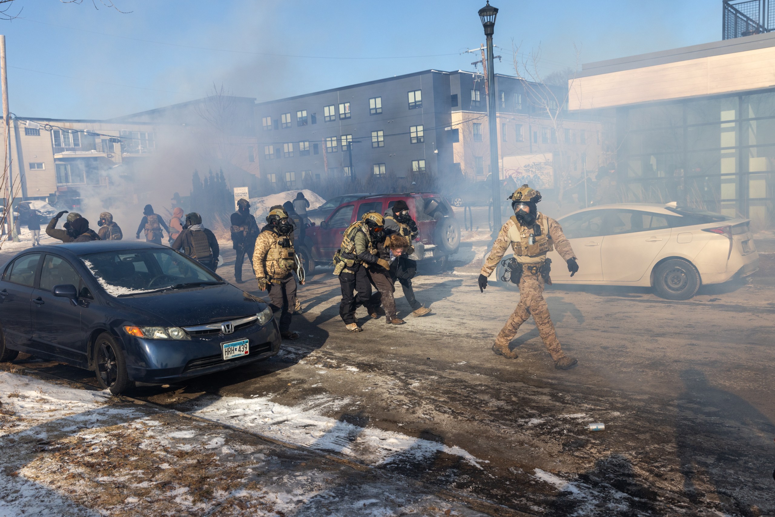 Several men in vests, helmets and gas masks walk through a haze of gas on an urban street. Two of them have a person in street clothes between them; the person is bent over double and their arms are being held. 