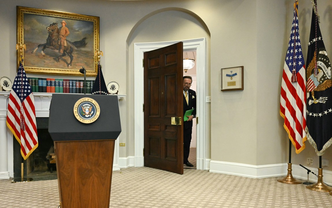 A man in a suit, carrying a green folder, looks through a large wooden door. On the other side is a room in the White House that has various flags, a painting, books and, at the forefront, a lectern with the presidential seal. 