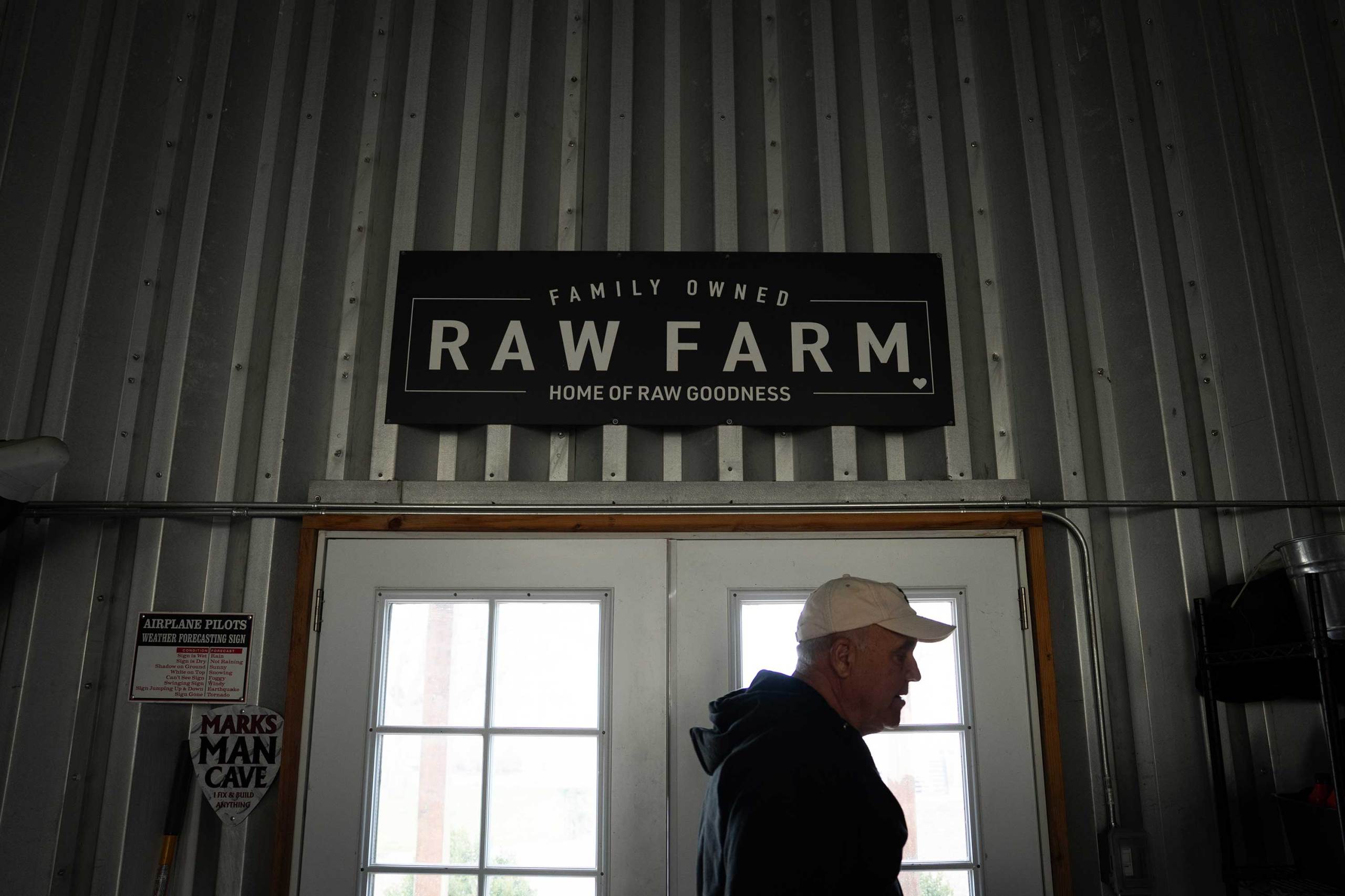 A man in a baseball cap walks past double glass doors inside a dimly lit building with corrugated metal walls. Above the doors hangs a large Raw Farm sign.