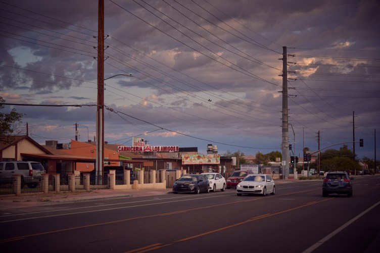 A suburban street flanked by large electrical poles supporting many rows of wires. Above them is a cloudy blue sky.