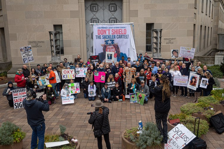 Several dozen protesters gather in front of news cameras. They hold numerous signs and stand in front of a large banner that reads “Don’t Shield the Sackler Cartel.” Behind them is a government building with “Department of Justice” written on it.