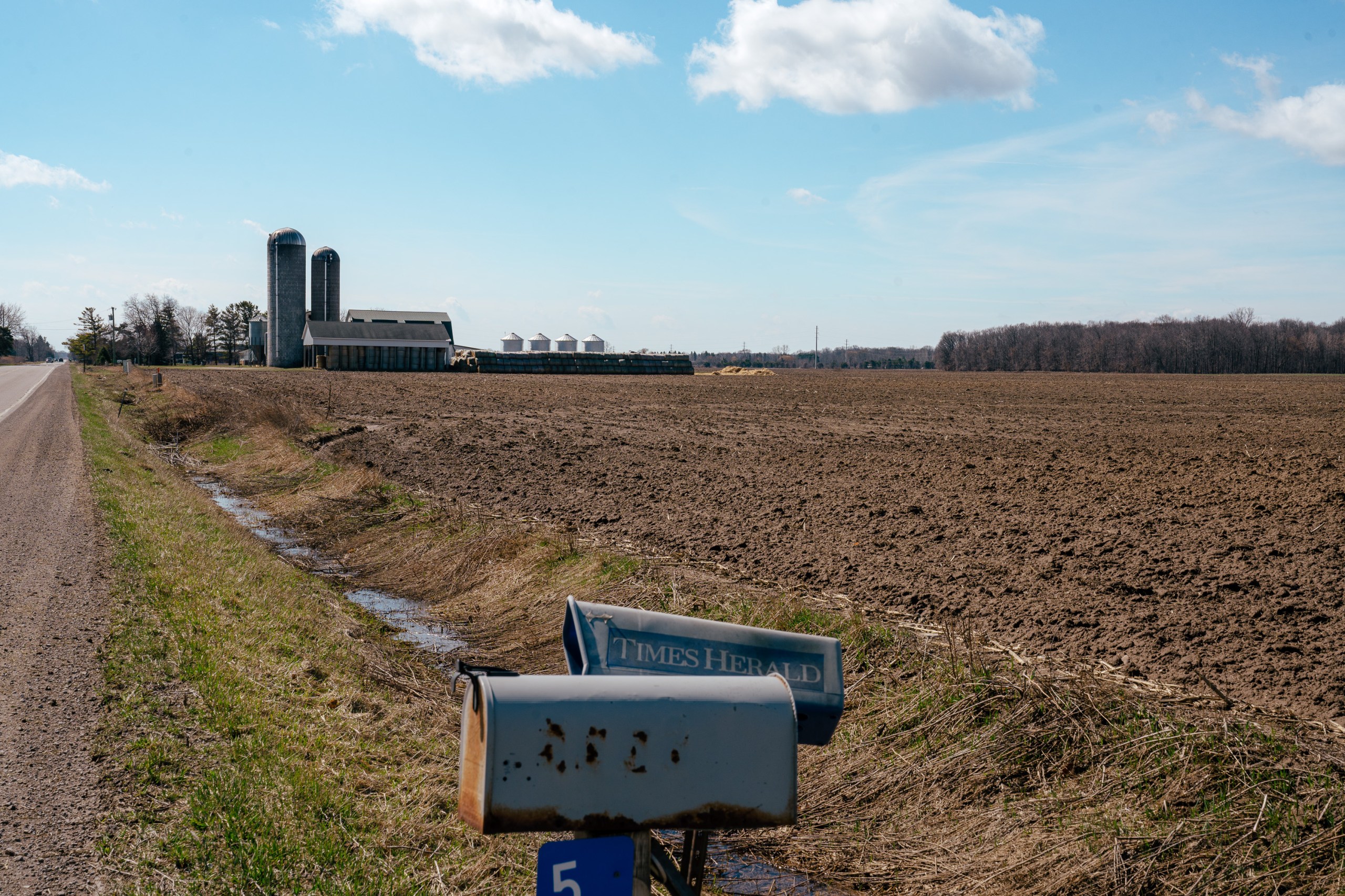 A rusty mailbox and a newspaper delivery box stand near a rural road. In the background is a large, recently tilled field, a large barn and two silos.