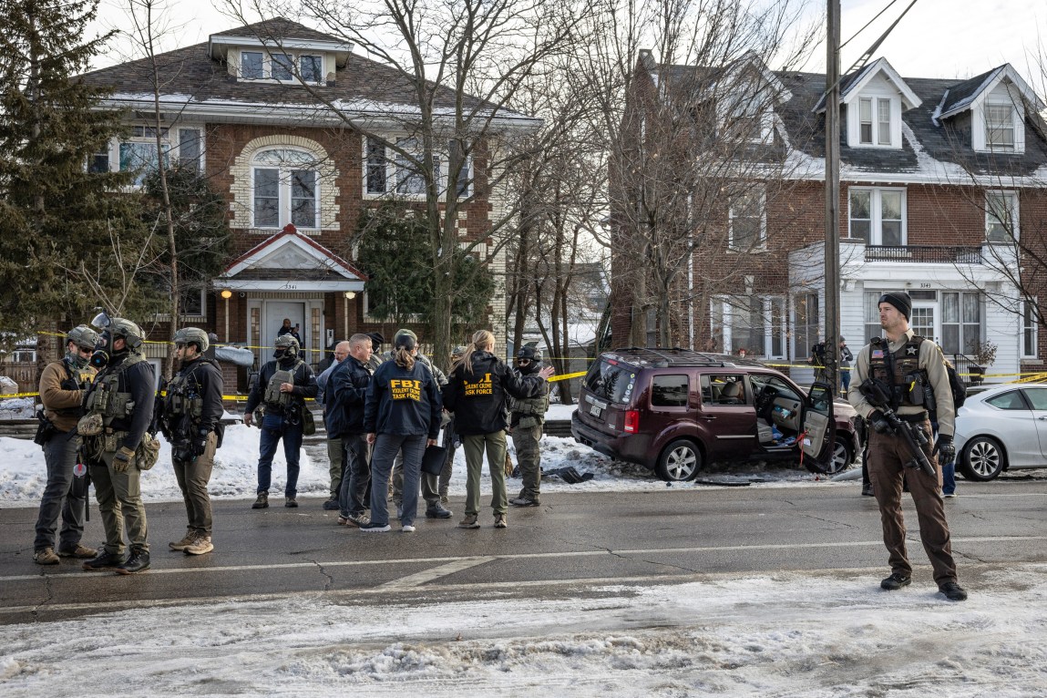 Groups of heavily armed law enforcement officials, including two with windbreakers that say “FBI,” stand in a residential street near a maroon SUV that has crashed into a utility pole.