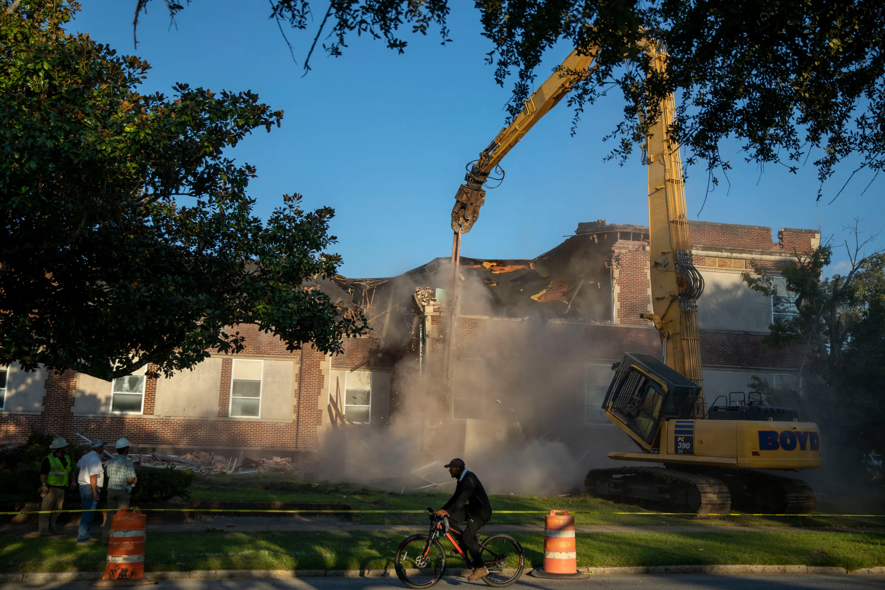 An excavator tears down a big red-brick building that is enveloped in dust. A man bikes by in the foreground.