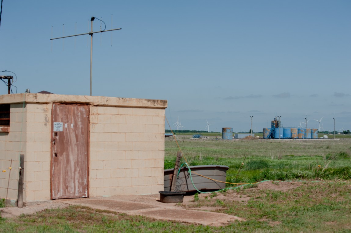 A small, decrepit building is on the left side of a photograph. On the right side, behind the building in a grassy field, is a row of blue industrial structures.