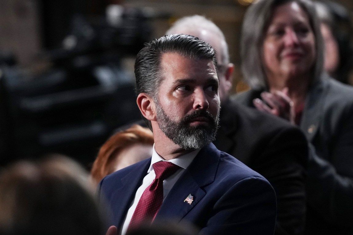 A man in a suit and tie, wearing an American flag lapel pin, looks to his left.