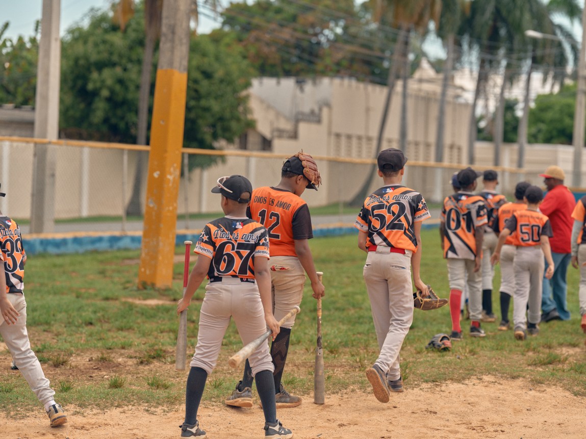 A group of young baseball players in orange, black and white baseball uniforms walk across a dirt field.