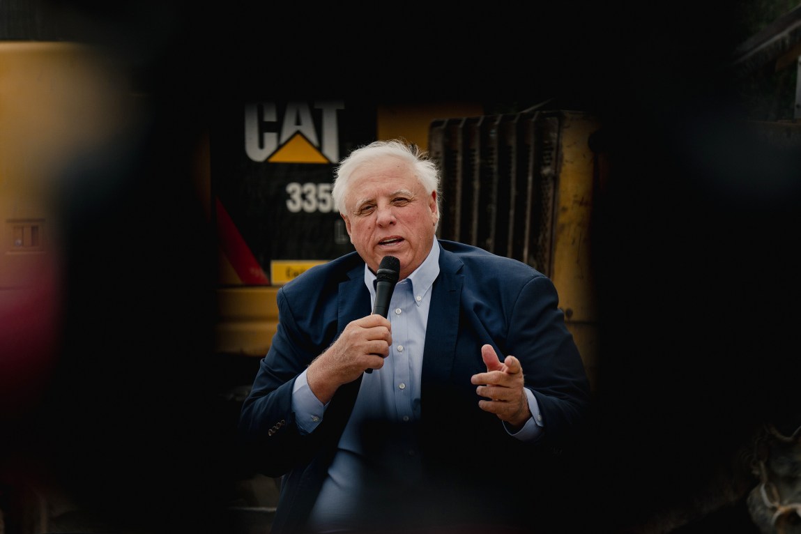 A man with gray hair, wearing a suit jacket, points with his left hand and speaks into a microphone. Behind him is construction machinery.