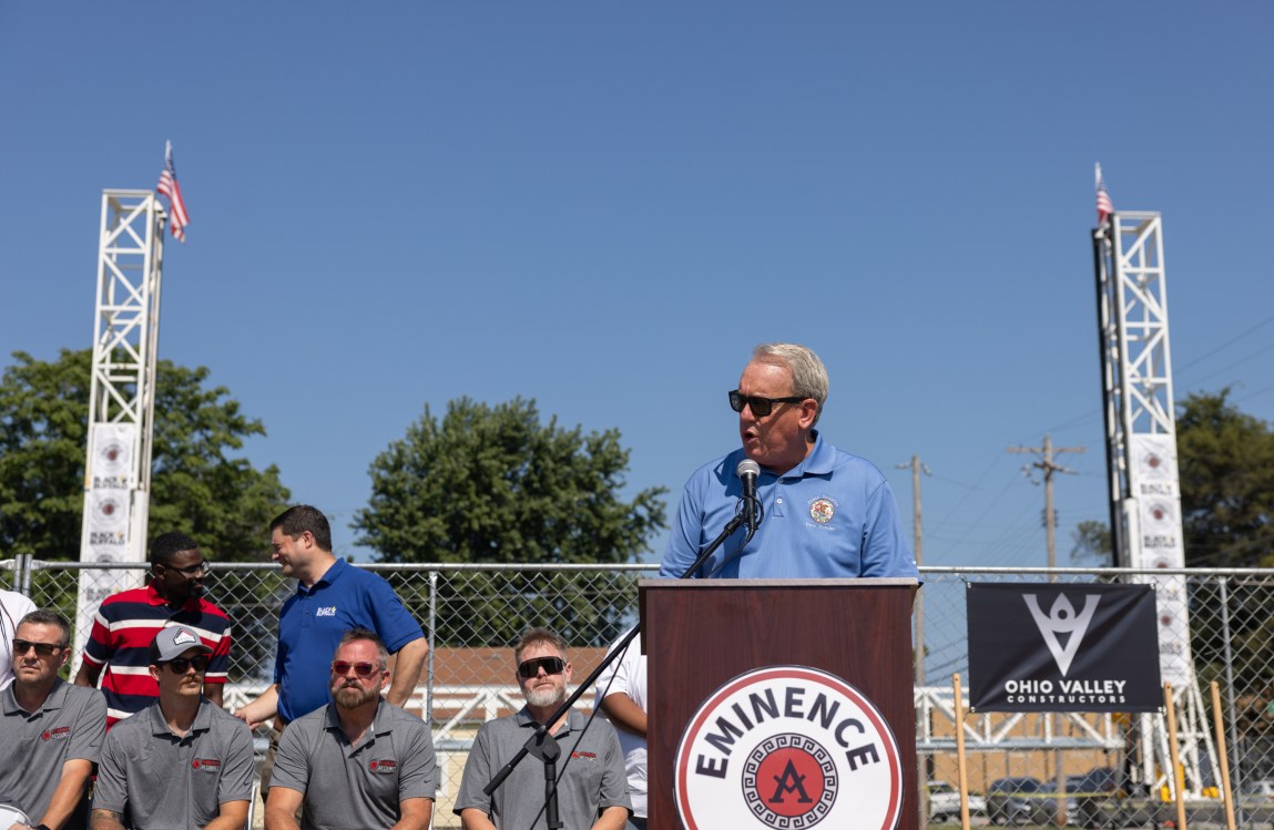 A man stands at a podium speaking into a microphone. Seated in a row next to him are men wearing gray shirts. Behind them are two tower-like structures, part of a huge 3D printer.