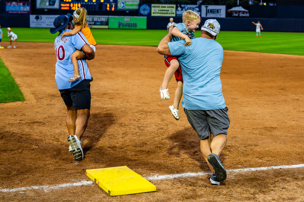 A pair of dualing fathers pick up their youngest children on first base as they race around the diamond to collect there children before reaching home during the fourth inning. The two dad's hilariously "forgot" their middle child on second base.
