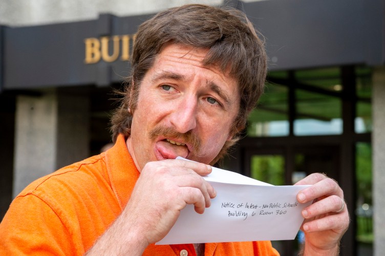 A close-up of a man licking the edge of an envelope flap. The envelope reads in handwritten letters: “Notice of Intent - Non Public Schools, Building 6 Room 360.”
