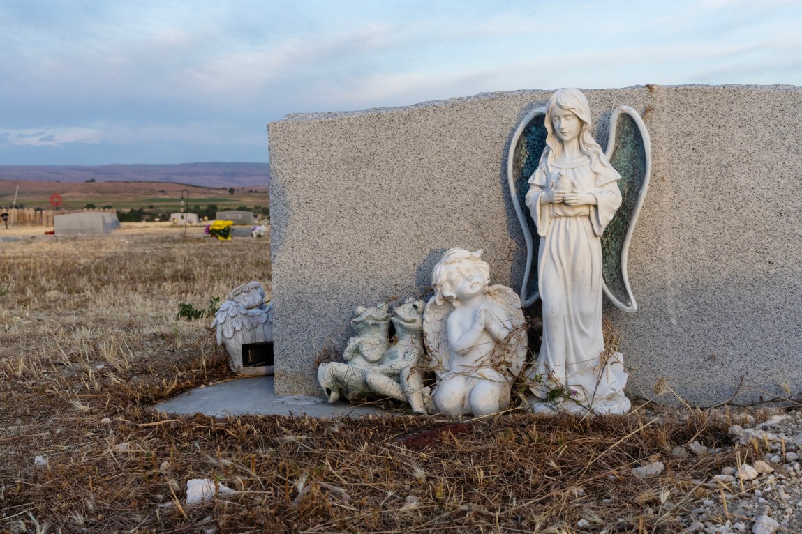 A gravestone with various statues: two frogs smiling, a cherub praying and a childlike angel. Brown grass stretches out behind the gravestone and mountains and a blue sky can be seen in the distance.
