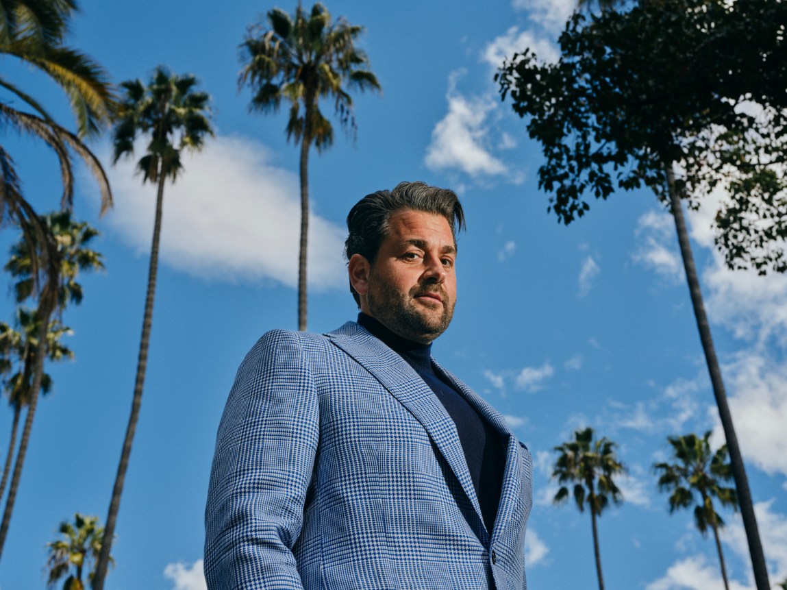 A man wearing a blue plaid suit and turtleneck looks down at the camera. Behind him are a blue sky and palm trees.