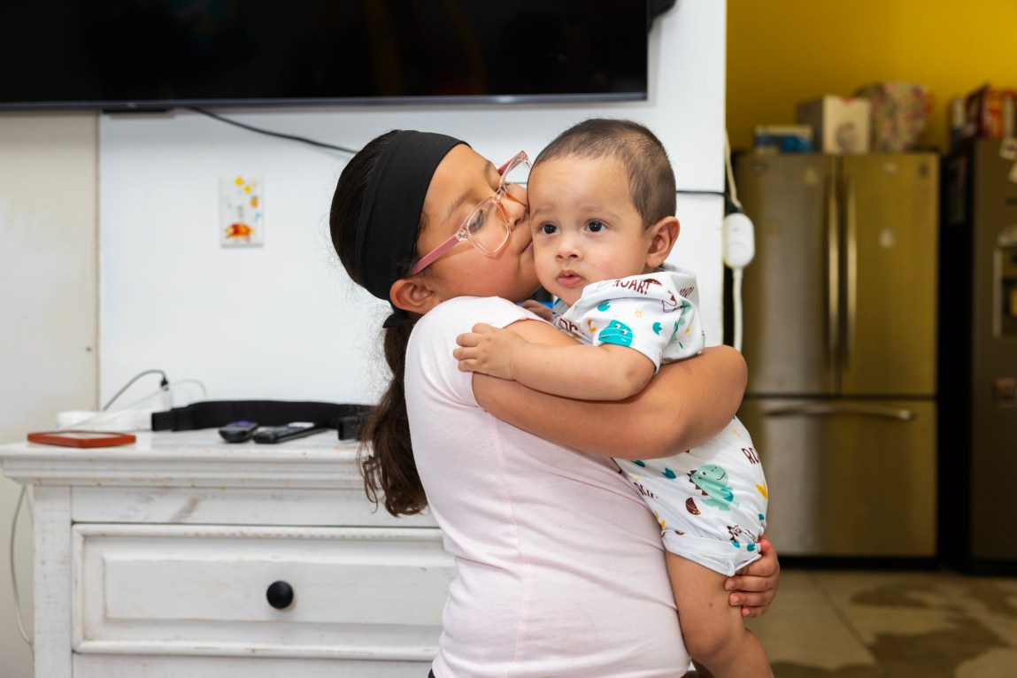 A child wearing a black headband, pink glasses and a pink shirt holds a baby wearing a white onesie with a dinosaur pattern. They are standing in front of a television and a white table. Behind them is a room with a silver refrigerator bathed in yellow light.