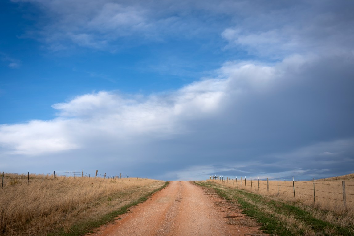 A wide, scenic shot of a dirt road cresting a hill, lined on both sides by wire fencing and dry grass, under a dramatic, cloudy blue sky.