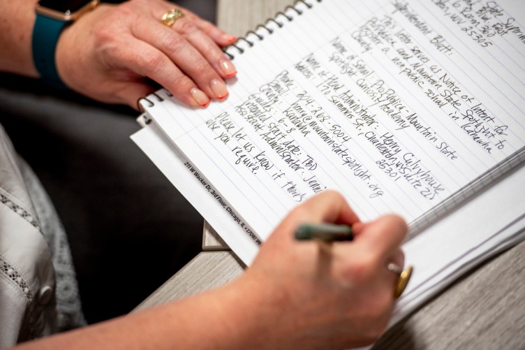 A close-up of a notebook with handwritten words. Hands hold the edge of the notebook and a pen.