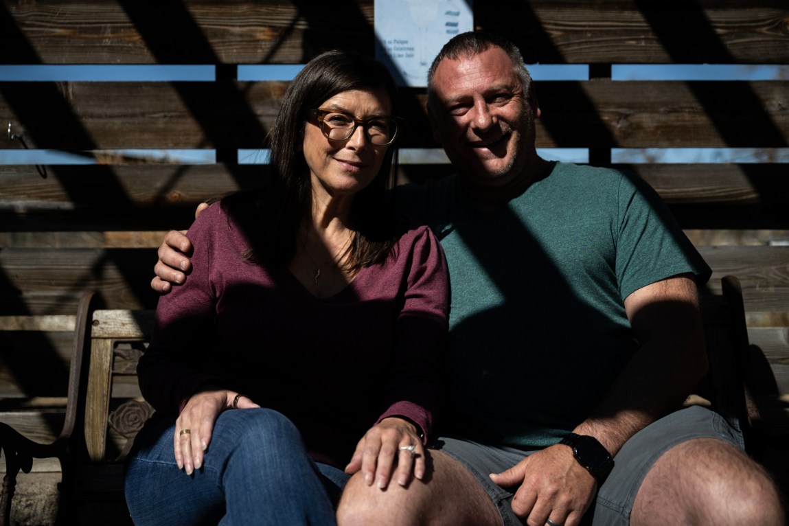 A smiling woman and a man sit next to each other on a wooden bench in front of a wooden fence. The man has his arm around the woman and the woman has her hand on his knee. Diagonal lines of shadow fall across their bodies.