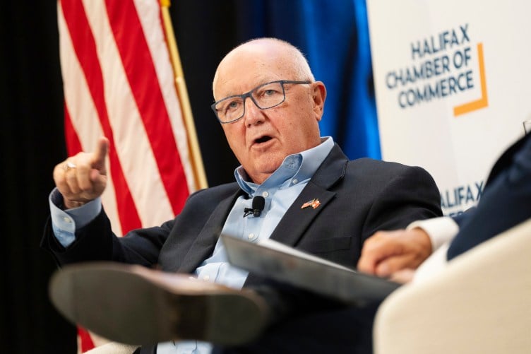 An older man with a bald head, wearing glasses and a dark blue suit, points while speaking on a stage with an American flag on a pole behind him.